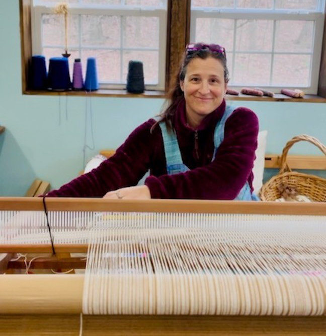 A woman sits at a weaving machine, smiling.